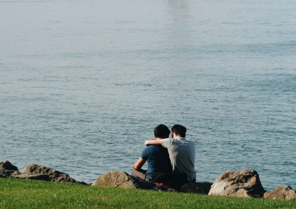 Zwei Männer sitzen am Wasser, einer legt den Arm um den anderen – Therapie für schwule Männer Berlin